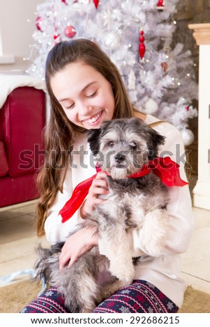 Young girl holding a Puppy at Christmas Time. She is smiling and the Puppy has a red ribbon around its neck.