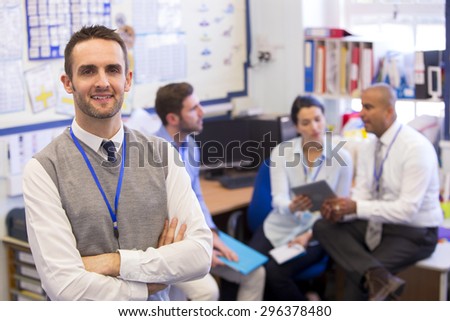 School teachers gather in a small school office for a chat. They look happy. The focus is on the man who stands in the foreground with the others sitting out of focus behind. He smiles at the camera.