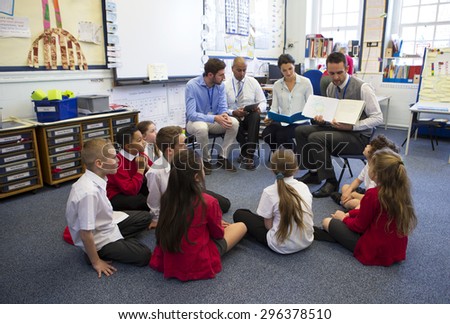 A group of children sit on the floor cross legged, listening to a group of teachers read a story.