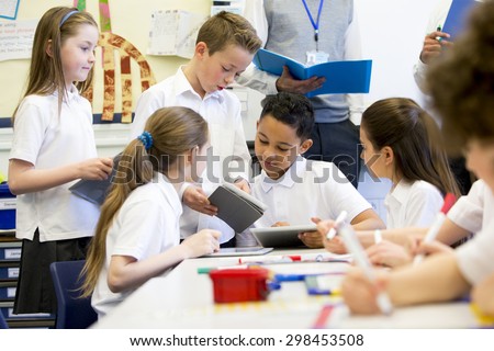 A group of school children can be seen working on digital tablets and whiteboards, they are all working happily. Two unrecognizable teachers can be seen in the background.