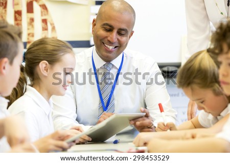 A male teacher sits supervising a group of children who are working on whiteboards and digital tablets.