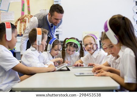 Group of children wearing colorful wireless headsets while working on digital tablets, the teacher can be seen supervising the students in the classroom