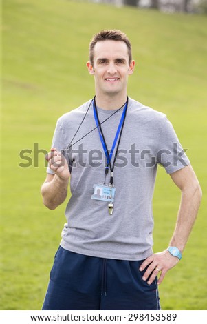 A male sports teacher stands outdoors on a field, he is holding a whistle.