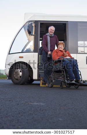 Elderly couple smiling for the camera as they get off the bus. The man is pushing his wheelchair bound wife.