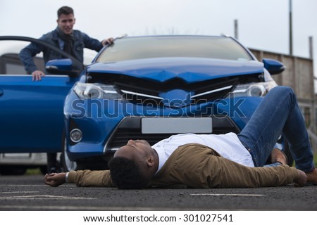 A man is lying in front of a damaged car which has just hit him. The responsible driver is standing at the door of the car, looking at him with a worried expression.