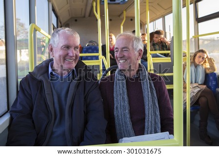 Two senior men smiling and talking on the bus. There are other people sitting on the bus in the background.