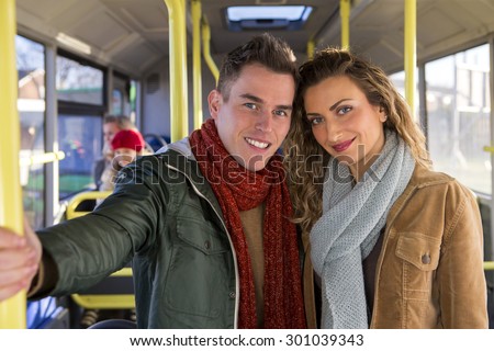 Young couple posing on a bus for the camera. They are standing up and there are people sitting on the bus in the background.