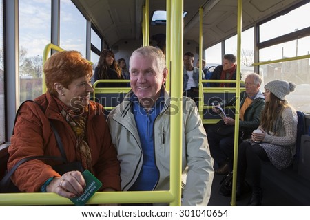 Senior couple travelling on the bus. There are other people sat on the bus who are in the background.