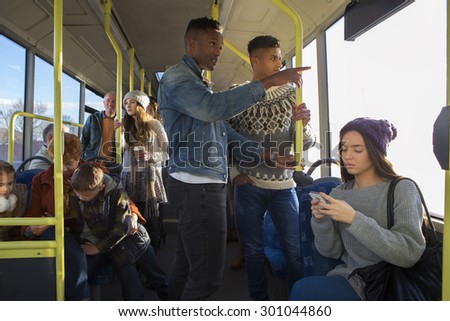 Two men are standing on a bus. There are people around them talking and using technology.