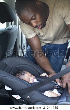 A father putting his newborn daughter into her car seat in the car. He is wearing casual clothing and looking at his daughter.