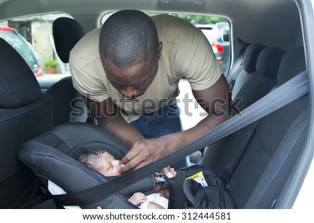 A father putting his newborn daughter into her car seat in the car. He is wearing casual clothing and looking at his daughter.
