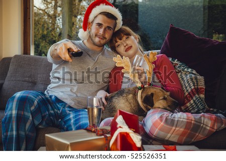 Young couple at home with their pet dog at Christmas time. They are all cuddled up on the sofa at home, watching television.