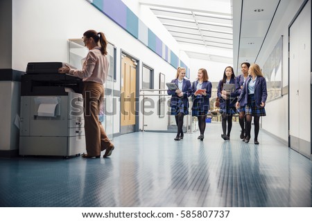 Group of teenage girls are walking down the school hall with books and laptops in their arms. They are talking and laughing as they walk and there is a female teacher using the printer.