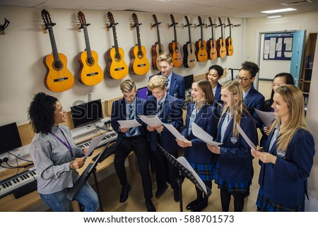 Class of teens are in their music lesson at school. There is a female teacher and the class are practicing choir with her.