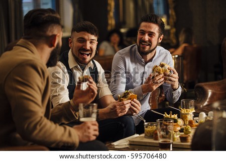 Three men are sitting together in a bar/restaurant lounge. They are laughing and talking while enjoying burgers and beer.