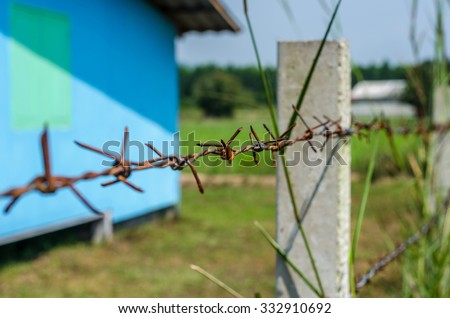 Rusty Barbed wire fence selective focus with blur overgrown grass and blue house background; countryside