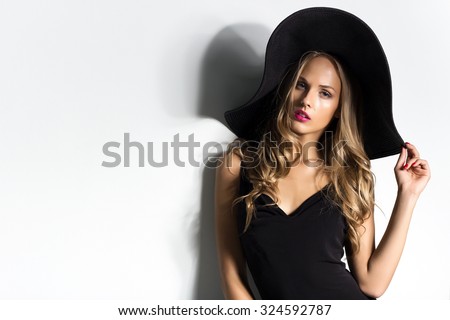 High fashion shot of blonde woman with curly hair in black hat and stylish elegant evening dress posing on white studio background.Red lips