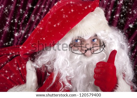 Blue-eyed Santa Claus carry a big red bag full with gifts and toys on his back and holding a thumbs up. Snow glitter backgrounds