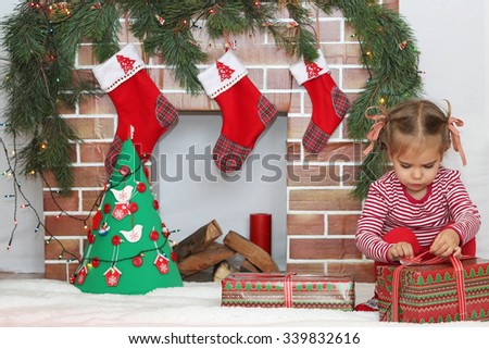 Pretty child girl opening a gift near Christmas decorated fireplace, winter holiday family concept