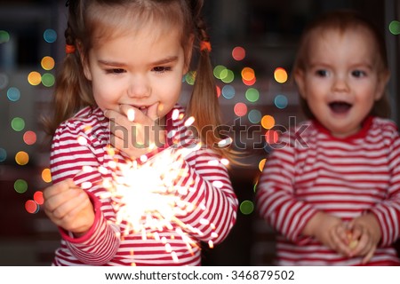 Handsome toddler boy and cute small girl in Santa hat hold burning sparkler and smile happily over defocused Christmas background