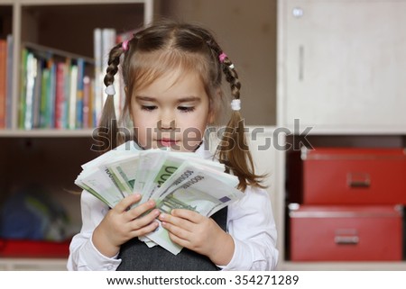 Inspired little child girl wearing in grey business style dress holds hundred Euro denominations (paper money) like a fan, indoor financial concept