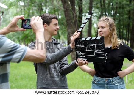 Teenage boy with realistic clapper cinema board ready to record a fun video with student girl in the park, a senior man holding smartphone to make a video, education and technology, outdoor summer