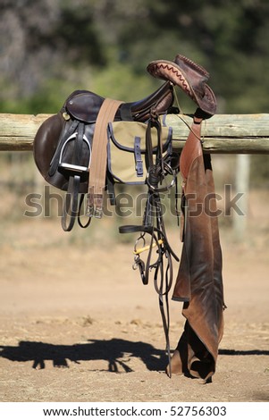 Riding tack, including leather hat, in full sunlight, displayed over a pole fence.