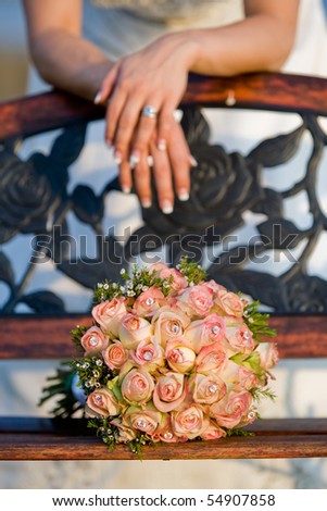 A vertical photo of a rose bouquet resting on a wooden bench, with a caucasian bride leaning behind it