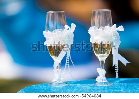 Two champagne glasses with white ribbons standing outside on a table with a blue table cloth