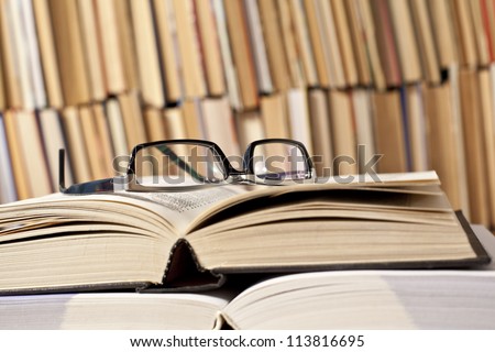 open and closed books and black glasses on  table front of a full bookshelf September 1st - the day of knowledge symbol of vision loss and eye strain from long reading