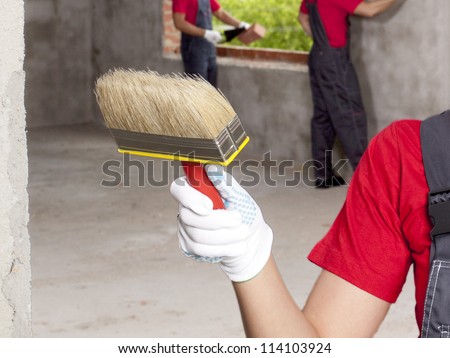 woman\'s hand in glove with brush paints a reinforced concrete wall - the background is out of focus - two men with bricks and masonry trowel