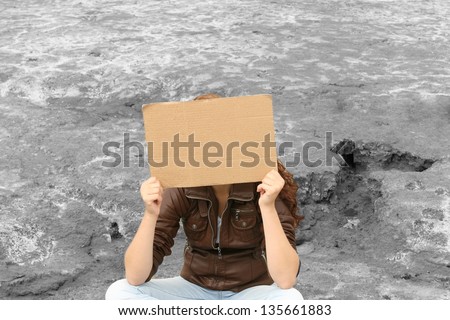 Abandoned teenage girl sitting outdoor on grey land and covers his face empty blank cardboard sign in her hands. Young girl looking sad and depressed. Neglected teens. Beggar and tramp. Unemployment.