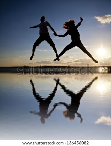 Silhouettes of couple jumping on sunset background with reflection on water