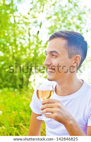 Young happy smiling brunette man with white wine, outdoors on green grass and trees background