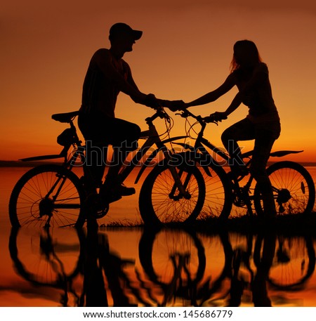 silhouettes of couples in love ride bicycles along the beach with reflection on water  Copy space for inscription