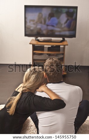 girl sits and rests on a strong sholder for a Man on the floor on TV background - couple embracing with each other