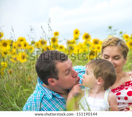 Copy space for inscription Family (mother, father, daughter) in sunflower field daughter asks dad to kiss her and shows a finger on her cheek