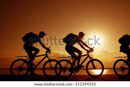 Image of sporty company three friends on bicycles outdoors against sunset. Silhouette A lot phases of motion of a single cyclist along the shoreline coast Reflection on water Space for inscription