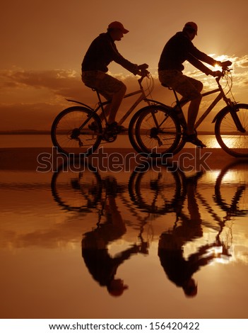 Image of sporty one couple - two friends on bicycles outdoors against sunset. Silhouette A lot phases of motion single cyclist along the shoreline coast Reflection on water Space for inscription