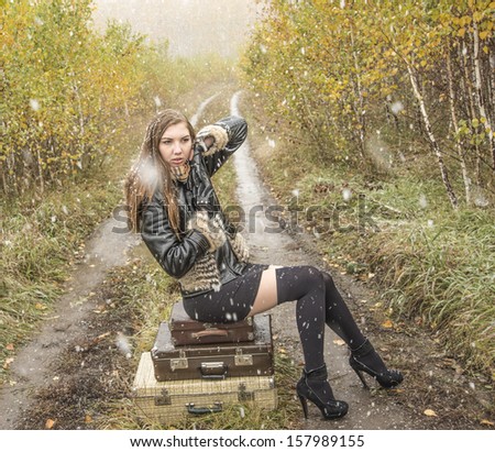 Young alone slim girl in a black leather jacket with fur collar sitting on the road in a forest in an old retro suitcase  on snowy background of the autumn snowfall