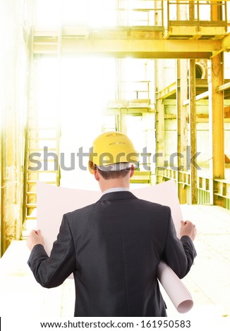 back view worker or engineer with yellow helmet for workers security holding in hands paper plan on the background of a new metal industrial brown building in perspective