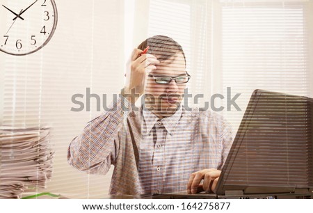 Young businessman in glasses hardworking and  looking at screen black laptop at desk in his office on background gray wall with clock Vertical