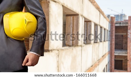 adult engineer or inspector hand holding yellow plastic helmet for workers security over a lot gray concrete windows and red bricks building background