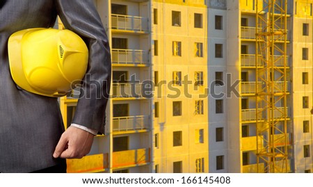 adult engineer or inspector hand holding yellow plastic helmet for workers security over multistories house at sunset light background No face Copy space for inscription