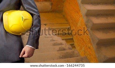 adult engineer or inspector hand holding yellow plastic helmet for workers security over gray concrete ladder in new building background