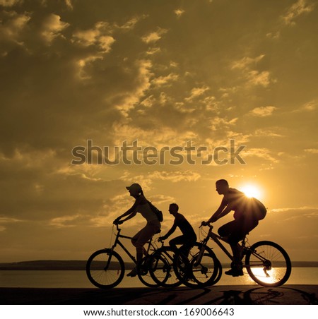 Image of sporty company three friends on bicycles outdoors against sunset. Silhouette motion of 3 cyclist along the shoreline coast and cloudy sunset sky Space for inscription