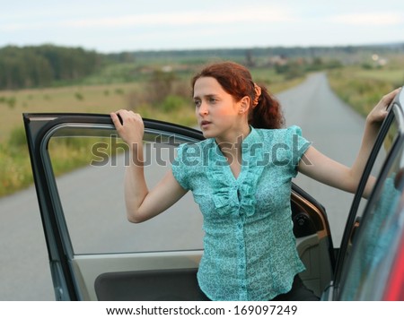 Young joyous beauty redhead long hair woman looking at opened car window keep door on asphalt road and blue cloudy sky background in perspective