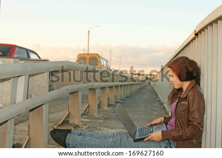 young Smiling beautiful woman with long red-head hair using black wireless laptop on a countryside road with cars wait your yellow bus on blue cloudy sky background Idea GPS navigator