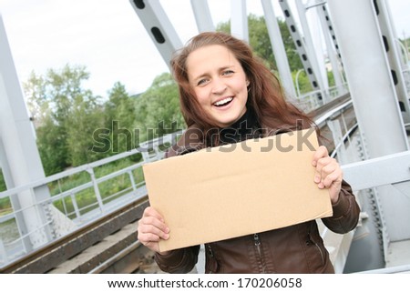Portrait of Redhead hair young Girl in brown leather jacket with empty cartoon cardboard sign on the background of green summer forest , blue cloudy sky and metal railway station bridge girders