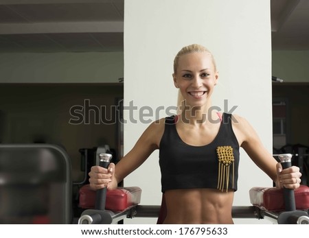 Fitness Woman with smiling face. Close-up portrait of young adult blond girl with long hair holding simulator for swing press on dark green wall background